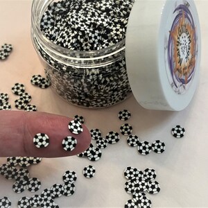 May include: A clear jar filled with black and white soccer ball shaped glitter. The jar is on a pink surface with some of the glitter scattered around it.