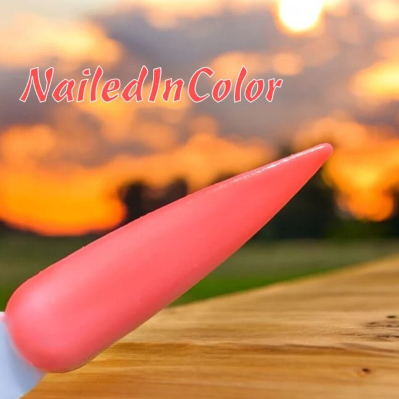 May include: A close-up of a bright pink nail polish tip, with a blurred sunset background and the text "Nailed In Color".