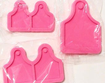 May include: Three bright pink silicone molds for crafting, each individually wrapped in clear plastic. Two molds feature a double-tag shape with circular holes at the top, while the third is a single, larger tag-shaped mold with a circular hole.