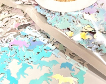 May include: A collection of iridescent, unicorn-shaped confetti pieces in shades of light blue, pink, and yellow. The small, flat, die-cut shapes are contained within a clear plastic bag, topped with a circular label featuring purple text.