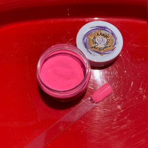 K&ouml;nnte beinhalten: Ein kleines Glas mit rosa Glitterpuder und einem durchsichtigen Deckel. Das Glas ist mit "Glitter-Magicco Make your magic even" beschriftet. Ein durchsichtiger Plastik-Applikator mit einer rosa Spitze liegt neben dem Glas.