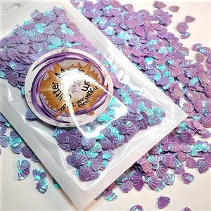 May include: A clear plastic bag filled with iridescent seashell-shaped glitter. The glitter is primarily purple with hints of teal and blue. A circular sticker with text is on the bag. The glitter is scattered around the bag.