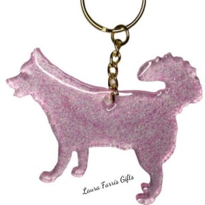 May include: A dog-shaped keychain made of translucent pink resin infused with fine glitter. The keychain features a gold-toned metal jump ring and chain attached to the top of the dog figure. The text Laura Farris Gifts is printed below.