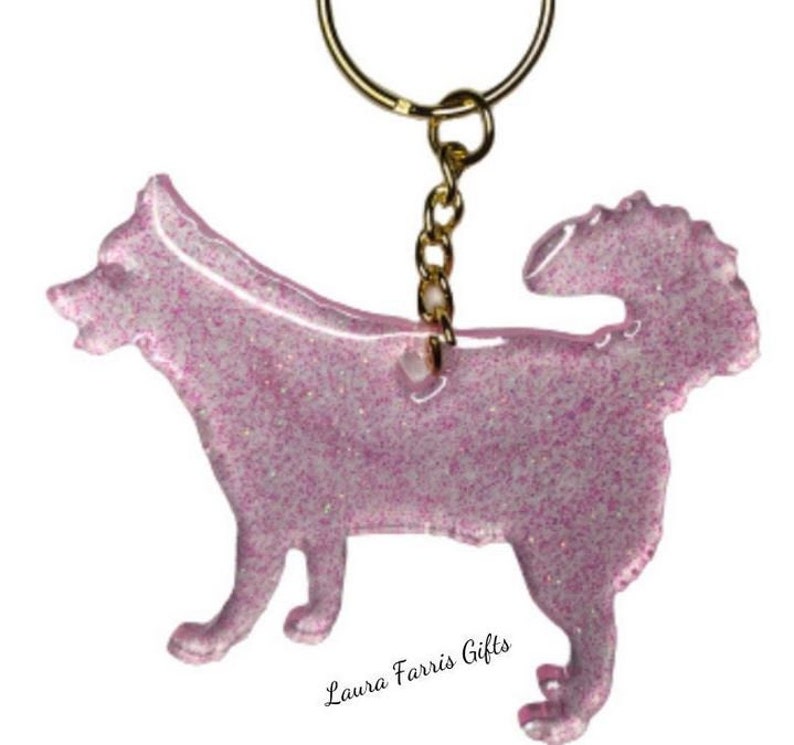 May include: A dog-shaped keychain made of translucent pink resin infused with fine glitter. The keychain features a gold-toned metal jump ring and chain attached to the top of the dog figure. The text Laura Farris Gifts is printed below.