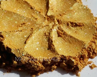 May include: A close-up of a pile of golden glitter with iridescent shimmer. The glitter is in a circular shape and has a textured surface. The text "MARYSGITTERMAGIC" is visible in the lower left corner of the image.