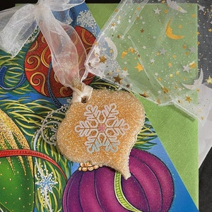 May include: A gold glitter heart-shaped ornament with a white snowflake design. The ornament is hanging from a white ribbon and is resting on a green and blue Christmas card with a purple and gold ornament.