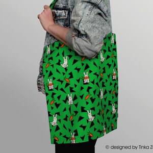 Pode incluir: Uma bolsa de tecido verde com um padrão de coelho branco e cenoura. A bolsa tem formas triangulares pretas em todo o design. O texto "designed by Tinka Zühlke" está na parte inferior da bolsa.
