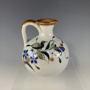May include: A small, round, white ceramic pitcher with a single handle and a brown-rimmed spout. The body is decorated with hand-painted black birds, green leaves, and small blue and orange flowers arranged in a flowing, organic pattern.