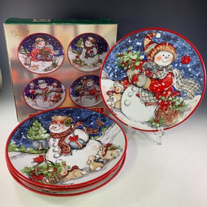 May include: A set of Christmas snowman plates with a red rim. The plates feature a snowman design with a red hat, scarf, and mittens, holding gifts and surrounded by snow and rabbits. The set includes a box with matching plates.