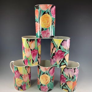 May include: Six ceramic mugs with floral designs. The mugs feature a black background with colorful flowers, including pink, yellow, and orange. The mugs are stacked in a pyramid shape.