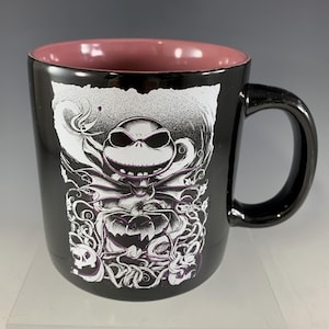 K&ouml;nnte beinhalten: Schwarze Keramik-Tasse mit einer wei&szlig;-lila Grafik von Jack Skellington aus The Nightmare Before Christmas. Die Tasse hat einen schwarzen Henkel und eine rosa Innenseite. Das Design umfasst einen K&uuml;rbis und wirbelnde Muster.