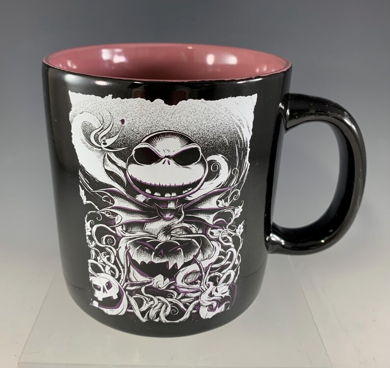 K&ouml;nnte beinhalten: Schwarze Keramik-Tasse mit einer wei&szlig;-lila Grafik von Jack Skellington aus The Nightmare Before Christmas. Die Tasse hat einen schwarzen Henkel und eine rosa Innenseite. Das Design umfasst einen K&uuml;rbis und wirbelnde Muster.
