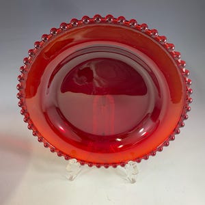 May include: A circular, translucent red glass plate featuring a decorative beaded edge. The plate has a smooth, reflective surface and a wide, flat rim that transitions into a shallow, concave center, showcasing a vibrant, uniform crimson color.