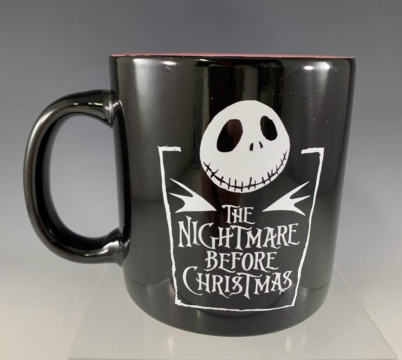 K&ouml;nnte beinhalten: Schwarze Keramik-Tasse mit der Figur Jack Skellington aus "The Nightmare Before Christmas". Die Tasse hat eine wei&szlig;e Grafik von Jacks Gesicht und dem Filmtitel. Der Innenrand ist rosa.