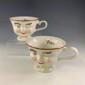 May include: Two white ceramic mugs with winking faces, heart-shaped handles, and bow ties. One mug has "Baileys" and "Yum" printed on it. The other mug has "Yum" printed on it. The mugs have brown trim and rosy cheeks.