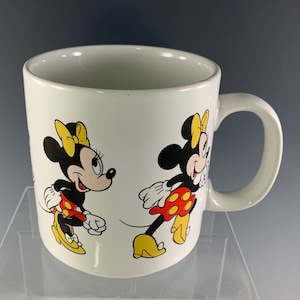 Puede incluir: Taza de cer&aacute;mica blanca con una imagen roja y amarilla de Minnie Mouse a cada lado. La taza tiene un asa negra.