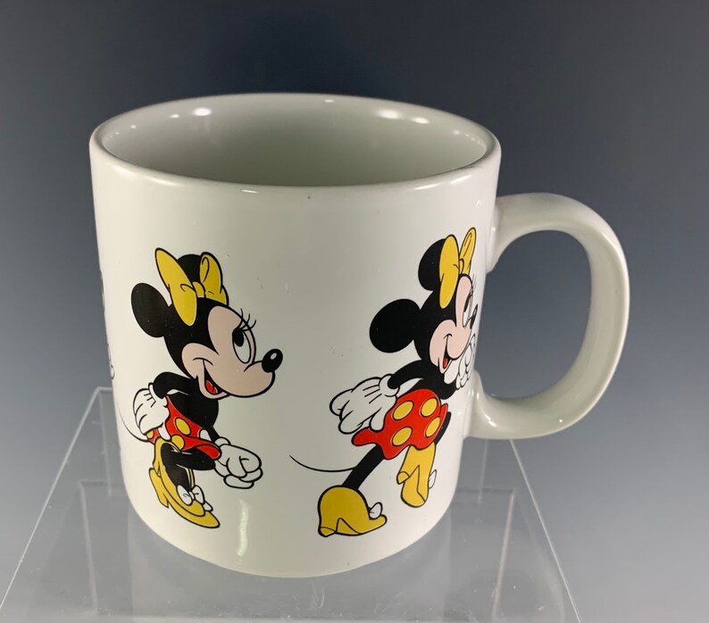 Puede incluir: Taza de cer&aacute;mica blanca con una imagen roja y amarilla de Minnie Mouse a cada lado. La taza tiene un asa negra.