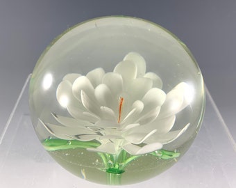 May include: A white glass flower encased in a clear glass sphere. The flower has a white center and green leaves.