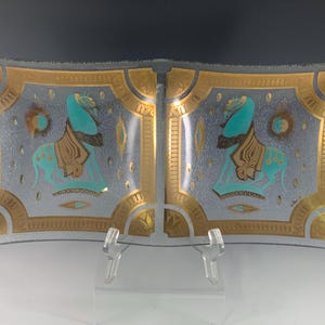 May include: Rectangular glass tray with a silver background and gold trim. The tray features two turquoise figures with gold accents. The design is symmetrical, with each figure facing the center. The tray is supported by a clear plastic stand.