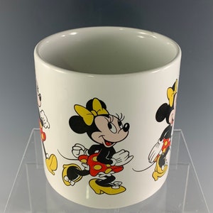 Puede incluir: Taza de cer&aacute;mica blanca con una ilustraci&oacute;n colorida de Minnie Mouse. La taza presenta a Minnie Mouse en tres poses diferentes, todas con su caracter&iacute;stico vestido rojo de lunares y zapatos amarillos.