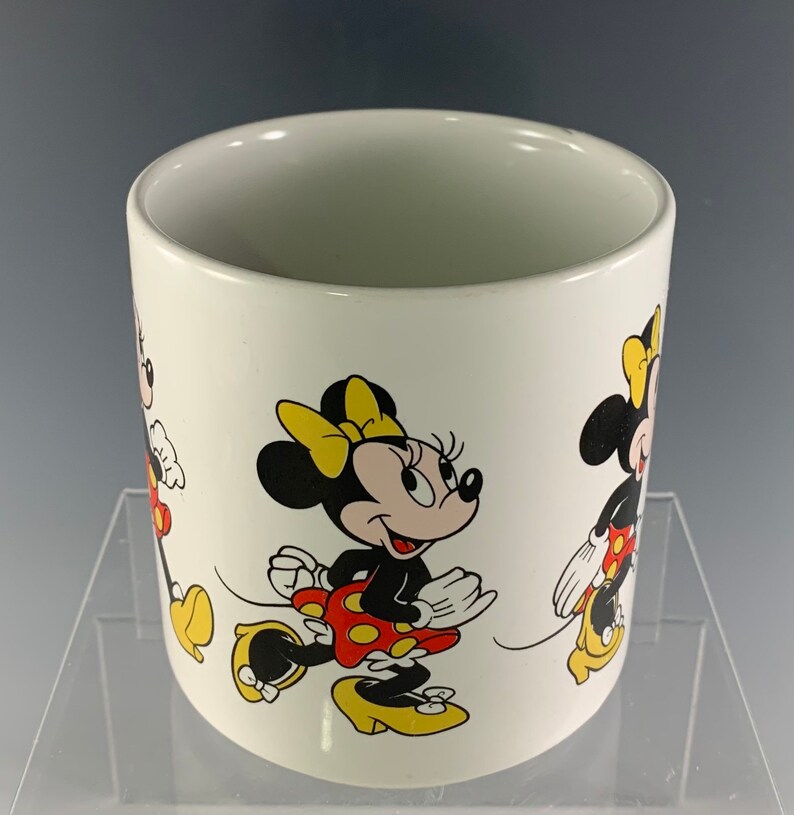 Puede incluir: Taza de cer&aacute;mica blanca con una ilustraci&oacute;n colorida de Minnie Mouse. La taza presenta a Minnie Mouse en tres poses diferentes, todas con su caracter&iacute;stico vestido rojo de lunares y zapatos amarillos.