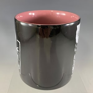 K&ouml;nnte beinhalten: Eine schwarze Keramiktasse mit rosa Innenraum. Die Tasse hat wei&szlig;en Text und ein Design auf der Au&szlig;enseite. Die Tasse ist zylindrisch und hat eine gl&auml;nzende Oberfl&auml;che. Die Tasse ist f&uuml;r hei&szlig;e oder kalte Getr&auml;nke geeignet.
