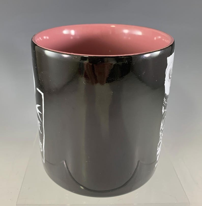 K&ouml;nnte beinhalten: Eine schwarze Keramiktasse mit rosa Innenraum. Die Tasse hat wei&szlig;en Text und ein Design auf der Au&szlig;enseite. Die Tasse ist zylindrisch und hat eine gl&auml;nzende Oberfl&auml;che. Die Tasse ist f&uuml;r hei&szlig;e oder kalte Getr&auml;nke geeignet.