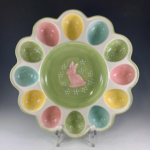 May include: A ceramic Easter egg serving platter. The platter is white with a scalloped edge and features pastel-colored egg-shaped compartments. The center has a green base with a pink bunny and white flower accents. The eggs are pink, yellow, green, and blue.