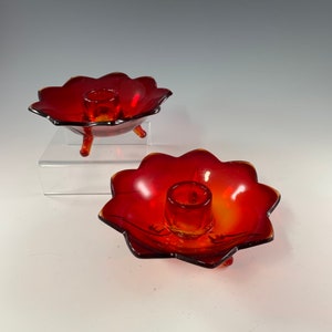 May include: Two vintage red glass candle holders. Each candle holder has a flower-shaped bowl with a central candle cup. One candle holder is elevated on three small legs. The glass has a gradient of red to orange.