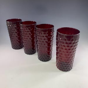 May include: Four tall, ruby-red drinking glasses. Each glass has a textured, dimpled surface and a cylindrical shape. The glasses are evenly spaced and stand upright, showcasing their rich color and design. Ideal for serving beverages.