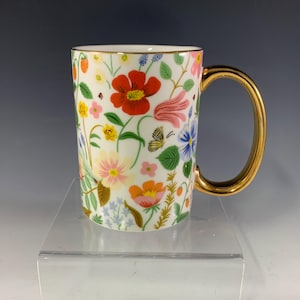 Może przedstawiać: Biały ceramiczny kubek ze złotym uchwytem i obramowaniem, ozdobiony kolorowym kwiatowym wzorem. Wz&oacute;r zawiera czerwone, r&oacute;żowe, ż&oacute;łte i niebieskie kwiaty, a także motyle i truskawki. Kubek ma około 13 cm wysokości.
