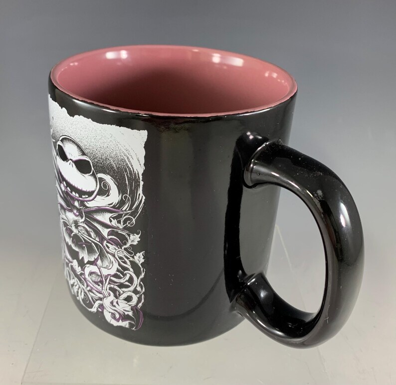 K&ouml;nnte beinhalten: Schwarze Keramik-Tasse mit gl&auml;nzendem Finish, mit einem Design von Jack Skellington aus The Nightmare Before Christmas. Der Innenraum ist staubrosa und der Griff ist schwarz. Das Design ist in Schwarz und Wei&szlig; mit lila Akzenten.