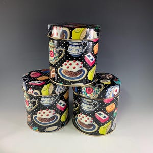 May include: Three cylindrical metal tea tins with lids, stacked. The tins are black with a white polka dot pattern and decorated with images of cakes, teacups, and teapots. The tins have gold rims.