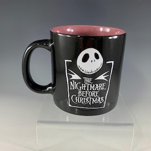 K&ouml;nnte beinhalten: Schwarze Keramiktasse mit rosa Innenraum. Die Tasse zeigt eine wei&szlig;e Grafik von Jack Skellingtons Gesicht und den Text "The Nightmare Before Christmas". Die Tasse hat einen schwarzen Griff.