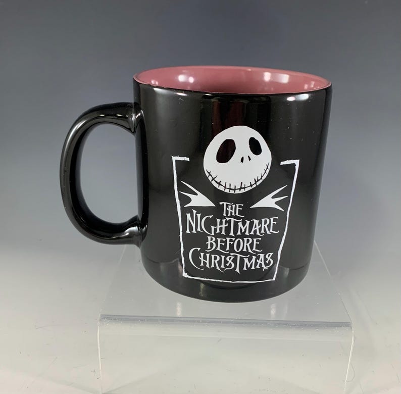 K&ouml;nnte beinhalten: Schwarze Keramiktasse mit rosa Innenraum. Die Tasse zeigt eine wei&szlig;e Grafik von Jack Skellingtons Gesicht und den Text "The Nightmare Before Christmas". Die Tasse hat einen schwarzen Griff.