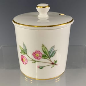 May include: White ceramic jar with lid, featuring a floral design of pink flowers and green leaves. The jar and lid are trimmed with gold accents. The lid has a small knob and a spoon rest.