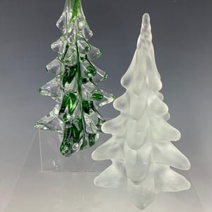 May include: Two glass tree figurines. One tree is made of clear glass with deep green internal coloring and a smooth, glossy finish. The second tree is made of frosted, translucent white glass with a matte, textured surface finish.