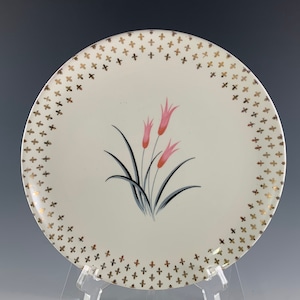 May include: A white ceramic plate with a gold floral pattern around the edge. The center of the plate features a pink and black floral design.