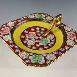 May include: A square, red and yellow ceramic dish with a gold handle. The dish is decorated with a floral pattern in shades of pink, green, white, and blue.