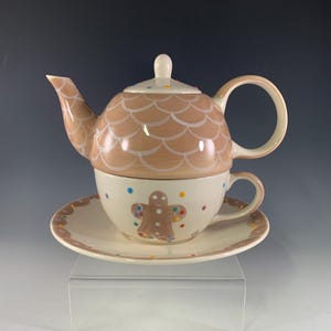 May include: A ceramic tea set featuring a teapot, teacup, and saucer. The teapot has a brown, scale-like pattern and a white handle. The teacup and saucer are off-white with a gingerbread man design and colorful polka dots. A charming holiday-themed tea set.