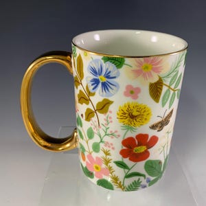 Może przedstawiać: Biały ceramiczny kubek ze złotym uchwytem i obramowaniem, ozdobiony kolorowym kwiatowym wzorem. Projekt zawiera niebieskie, r&oacute;żowe, ż&oacute;łte i czerwone kwiaty, a także zielone liście i motyla. Dekoracyjny kubek.