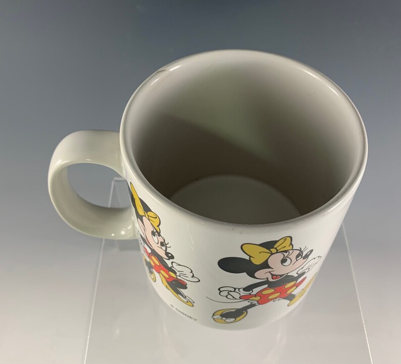 Puede incluir: Taza de cer&aacute;mica blanca con una imagen roja y amarilla de Minnie Mouse. La taza tiene un asa y la imagen de Minnie Mouse se repite en la taza.