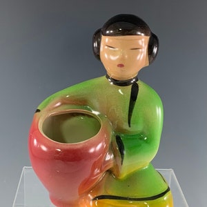May include: A ceramic planter shaped like a kneeling woman with a red and green glaze. The woman is holding a red planter pot in her hands.