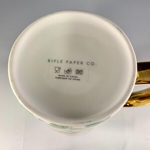 Może przedstawiać: Biały ceramiczny kubek ze złotym uchwytem i kwiatowym wzorem. Sp&oacute;d kubka zawiera tekst "RIFLE PAPER CO." i "MADE IN CHINA FABRIQUE EN CHINE". Kubek ma kolorowy kwiatowy wz&oacute;r z zielonymi, r&oacute;żowymi i niebieskimi akcentami.