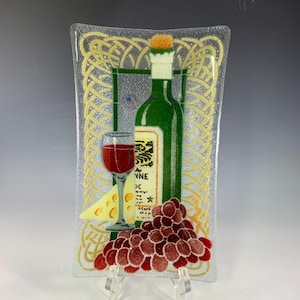 May include: Rectangular glass serving tray with a wine theme. The tray features a green wine bottle, a glass of red wine, a wedge of cheese, and a cluster of red grapes. The background has a gold Celtic knot design.