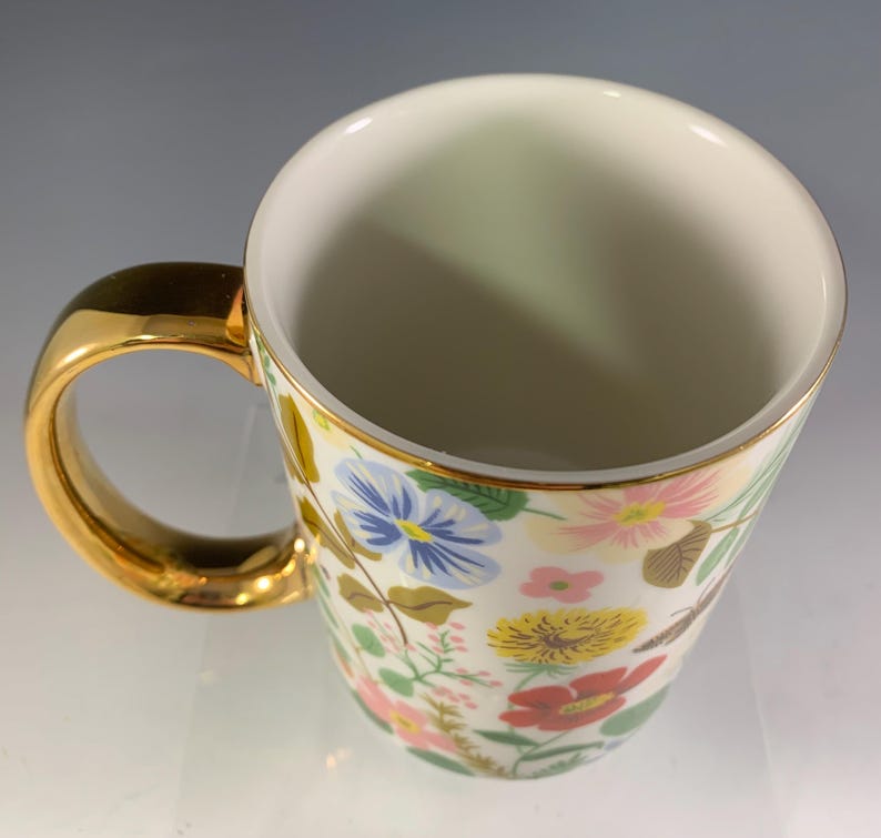 Może przedstawiać: Biały ceramiczny kubek ze złotym uchwytem i obramowaniem. Kubek jest ozdobiony kolorowym kwiatowym wzorem z czerwonymi, ż&oacute;łtymi, niebieskimi i r&oacute;żowymi kwiatami. Kubek jest pusty.