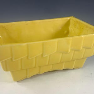 May include: A rectangular, yellow ceramic planter with a textured, layered design. The planter has a slightly curved base and a smooth interior. The yellow color is consistent throughout the piece, giving it a cohesive look.