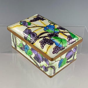 May include: A small rectangular box with a white background and a floral design. The flowers are purple, green, and blue, and the leaves are green. The box has a gold trim.