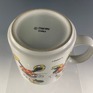 Puede incluir: Taza de cer&aacute;mica blanca con un contorno negro de Mickey Mouse y Minnie Mouse. La taza tiene un asa negra y el texto "&copy; Disney" y "KOREA" en la parte inferior.