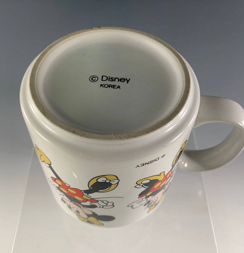 Puede incluir: Taza de cer&aacute;mica blanca con un contorno negro de Mickey Mouse y Minnie Mouse. La taza tiene un asa negra y el texto "&copy; Disney" y "KOREA" en la parte inferior.